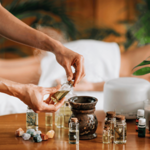 An image showing a person's hands as they prepare for an aromatherapy session, likely taken in a spa in Burnaby. The focus is on a small, clear bottle filled with yellow liquid that the hands are opening. There's a dark brown burner with a lit candle inside, creating a relaxing mood. Several small bottles with different oils and colorful crystals are scattered around the table, adding to the calm and natural vibe. Behind, there is a blurred white towel, suggesting a massage table for an aromatherapy massage. The atmosphere is serene, perfect for a healing and soothing experience in Burnaby.