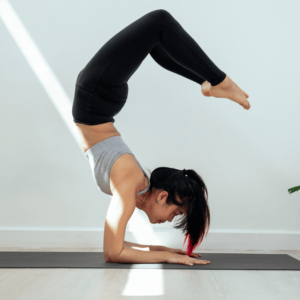 <br />
The image features a person practicing a yoga pose on a gray mat against a bright white wall. The individual is in an inverted pose, supporting their body weight with their forearms flat on the mat, their head tucked in, and legs bent at the knees, forming a sharp angle. They are wearing black leggings and a light gray tank top, highlighting the lines of their body against the minimalist background. The person's hair is tied back with a hint of red color showing, and a beam of sunlight cuts across the scene, adding contrast and depth to the image. This pose showcases flexibility, strength, and the focus required for such a balance, often associated with a healthy and active lifestyle.