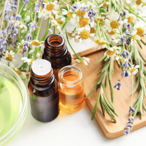 This image shows a bright and cheerful display of aromatherapy oils, perfect for someone looking for a relaxing massage in Burnaby. Three amber glass bottles with essential oils are on a bamboo wooden board. One bottle is open, revealing a clear, golden liquid. There's a mix of fresh lavender and white daisy flowers scattered around, with some lavender stems elegantly draping over the edge of the board. In the corner is a hint of a light green bowl, adding a splash of color to the scene. The whole setup is on a white surface, bathing in soft, natural light that highlights the vibrant colors of the flowers and the rich hues of the essential oils. This image captures the essence of a calming aromatherapy atmosphere.