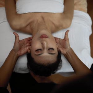 This photo captures a moment of relaxation during a neck massage, which might remind someone of the soothing touch they can expect at an aromatherapy massage in Burnaby. The picture shows a person lying on their back, with their head slightly tilted back, eyes closed in relaxation. They're wearing a white tube top, and their skin is glowing against the white sheets of the massage table. Two hands are gently cradling their head, one at the back of the neck and the other at the crown, suggesting the beginning of a head massage. The focus is on the person's serene expression and the hands of the therapist, conveying a sense of peace and expert care. The background is softly blurred, emphasizing the tranquil experience.