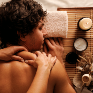 The image shows a person lying face down on a massage table, receiving a relaxing back massage that might remind you of a therapeutic experience in Burnaby. The person's back is bare, and the focus is on the hands of the therapist as they press into the shoulder muscles. A rolled-up white towel is placed under the person's forehead for comfort, and their curly hair is partly visible. To the side, we can see massage essentials: a lit candle, providing a soft glow and ambiance, a container of massage balm, and a small arrangement of dried plants, enhancing the feeling of a tranquil, natural setting. The warm tones in the picture suggest a cozy atmosphere, ideal for an aromatherapy massage.