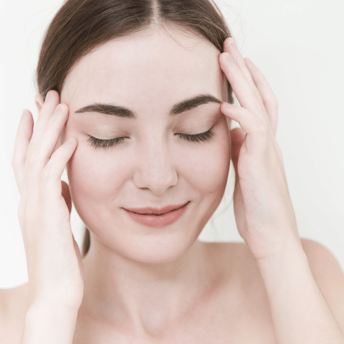 The image shows a close-up of a serene young woman with her eyes gently closed and a subtle smile on her lips, suggesting a moment of relaxation and stress relief, possibly after an enjoyable aromatherapy session in Burnaby. She's holding her hands up to her temples, lightly touching her face in a soothing manner. Her skin appears soft and clear, and she has neatly groomed eyebrows, natural makeup, and long eyelashes. The background is a plain, light color, emphasizing the calm and clean atmosphere. The woman's expression and gentle touch to her head convey a feeling of tranquility and self-care.