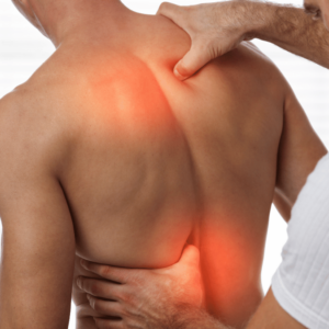 The image shows a close-up of a bare-backed person receiving a deep tissue massage from a therapist, aimed at relieving back pain, which could be a service found in a Burnaby massage clinic. The therapist's hands are applying pressure to the person's back, with the right hand on the upper back and the left around the lower back area. There's a red glow on the skin where the therapist's right hand is pressing, suggesting heat or a special light to highlight the area of focus and tension. The person's skin is smooth and the backdrop is neutral, drawing attention to the therapeutic touch and the area of discomfort. The image conveys a sense of relief and the healing touch of professional hands at work.