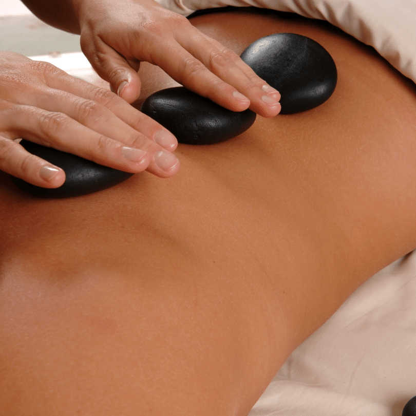 Close-up of a hot stone massage session showing the hands of a skilled therapist placing smooth, black basalt stones in perfect alignment down the client's spine. The warm stones are strategically positioned to target tense muscles, promoting deep relaxation and relief from muscular tension. The skin is glowing with a healthy sheen, indicating the gentle warmth of the stones, which are essential in this traditional therapeutic technique. This relaxing treatment is typical of what one might experience during a hot stone massage session designed to soothe and rejuvenate the body.