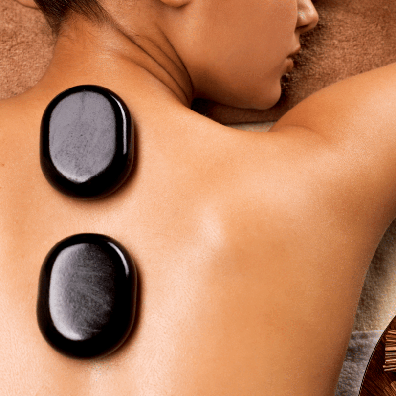 An overhead, close-up image showcasing a person lying face down receiving a hot stone massage, with two large, polished basalt stones resting on their back, along the shoulder blades. Initially, the stones appear to radiate therapeutic heat that deeply penetrates the muscles, thereby aiming to reduce stress and tension. Subsequently, the person's skin displays a warm, flushed tone indicative of increased circulation as a result of the hot stone application. Furthermore, this technique, integral to hot stone massage practices, is renowned for its ability to provide a deep sense of relaxation and wellness, traits highly sought after in massage therapy. Lastly, the composition of the image, with its focus on the stones and the recipient's relaxed posture, encapsulates the calm and healing environment typical of a hot stone massage experience.