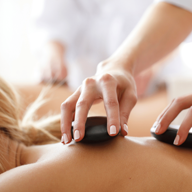 Image presents a serene hot stone massage in progress, with a focus on a massage therapist’s hands gently placing smooth, dark stones on the back of a reclining client. Initially, the therapist's hands, with neatly manicured nails, convey expertise and care as they strategically position the heated stones along the client's spine. Furthermore, the stones are shown as the central element, their dark silhouette against the client's light-toned skin illustrating the stark contrast and emphasizing the heat therapy in use. In addition, the background is intentionally blurred to draw attention to the precise action of the hot stone placement. This practice is part of the larger therapeutic hot stone massage technique, known for its relaxation and muscle tension relief benefits, and this image effectively encapsulates the peaceful ambiance that is synonymous with the experience.