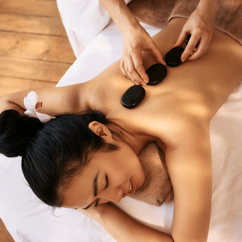 Top-down image capturing a peaceful moment during a hot stone massage, showcasing a content client with closed eyes and a serene expression. The client is lying face down on a white massage table, with a fluffy towel wrapped around the waist for comfort. Two large, smooth, black hot stones are placed with precision on either side of the client’s spine, around the mid-back area, by a massage therapist whose hands suggest a gentle, yet skillful touch. The warmth of the stones stands out as they rest against the client's radiant skin, which is meant to facilitate relaxation of muscles and promote deep tissue therapy. The relaxed ambiance is further accentuated by the wooden floor of the massage room and the delicate white flower placed beside the client’s head, symbolizing purity and tranquility. Overall, the image embodies the essence of a hot stone massage, which is intended to create a harmonious balance of body and mind.