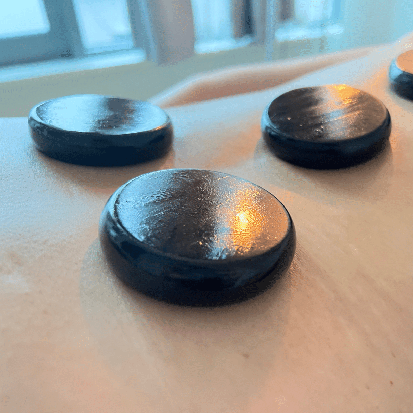 A close-up image depicting a trio of smooth, black hot stones used for a hot stone massage, carefully placed on a person’s back, glistening with a sheen of oil. The stones are in sharp focus in the foreground, emphasizing their rounded edges and the glossy texture that comes from the massage oil, reflecting a soft, warm light that suggests a soothing warmth. The background, blurred for emphasis on the stones, hints at a tranquil spa environment with soft lighting that creates a calming atmosphere. The stones are arranged to align with the spine, targeting key points along the back to facilitate muscle relaxation, improve circulation, and provide a deeply relaxing experience as part of a professional hot stone massage therapy session.