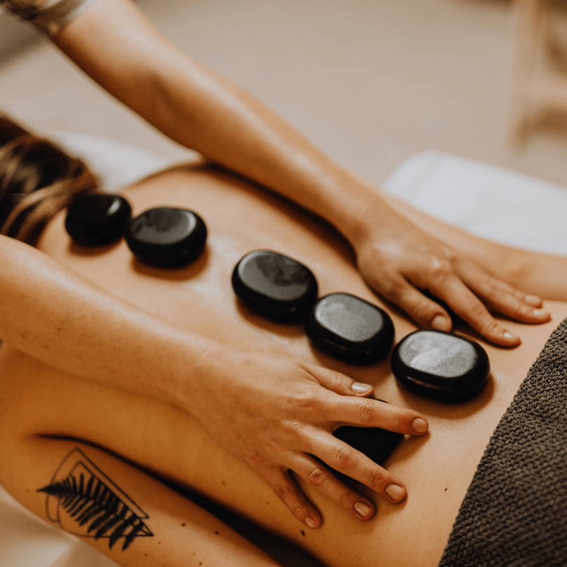 An intimate and warmly lit scene of a hot stone massage in progress, featuring a client lying face down on a massage table. The client’s skin is adorned with a tattoo of a fern on the left side, adding a personal touch to the image. Five smooth, volcanic stones are strategically placed along the client's spine, radiating therapeutic heat. A massage therapist's hands are seen in the act of gently pressing another stone against the client's lower back, with their fingers demonstrating a professional and gentle technique. The stones, matte black and perfectly rounded, stand out against the soft, neutral tones of the client's skin and the massage table, illustrating a tranquil and healing environment. This image encapsulates the essence of a hot stone massage, which is believed to relax the muscles, alleviate stress, and harmonize the body's energy.