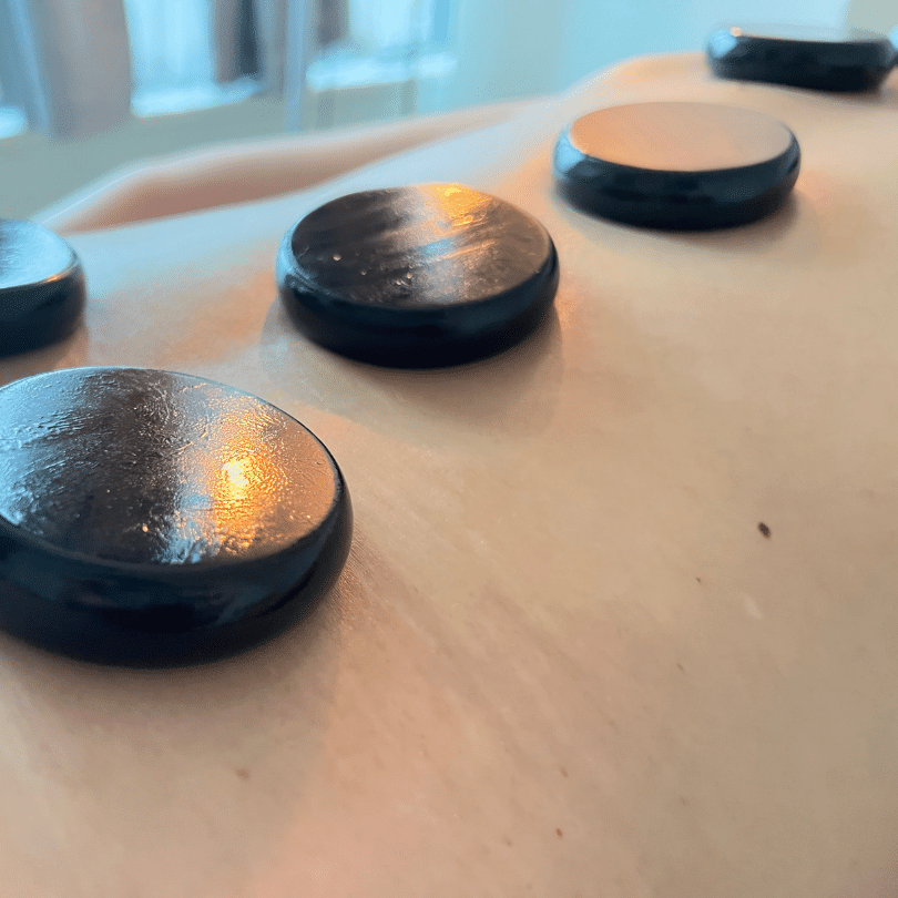 A close-up photo captures the essence of a hot stone massage with three circular, smooth basalt stones placed directly on the client’s back, their surfaces reflecting the soft, ambient light of the room. The stones, renowned for their heat retention properties, are placed along the spine, which is a common technique in hot stone massage therapy to encourage relaxation and release of muscle tension. The warmth from the stones is visible through the slight glistening of the massage oils on their black surface, suggesting the soothing temperature that is central to the treatment. The gentle gradient of light across the image accentuates the curvature of the client's back and the rounded edges of the stones, providing a sense of the tranquil atmosphere and therapeutic touch involved in a hot stone massage session.