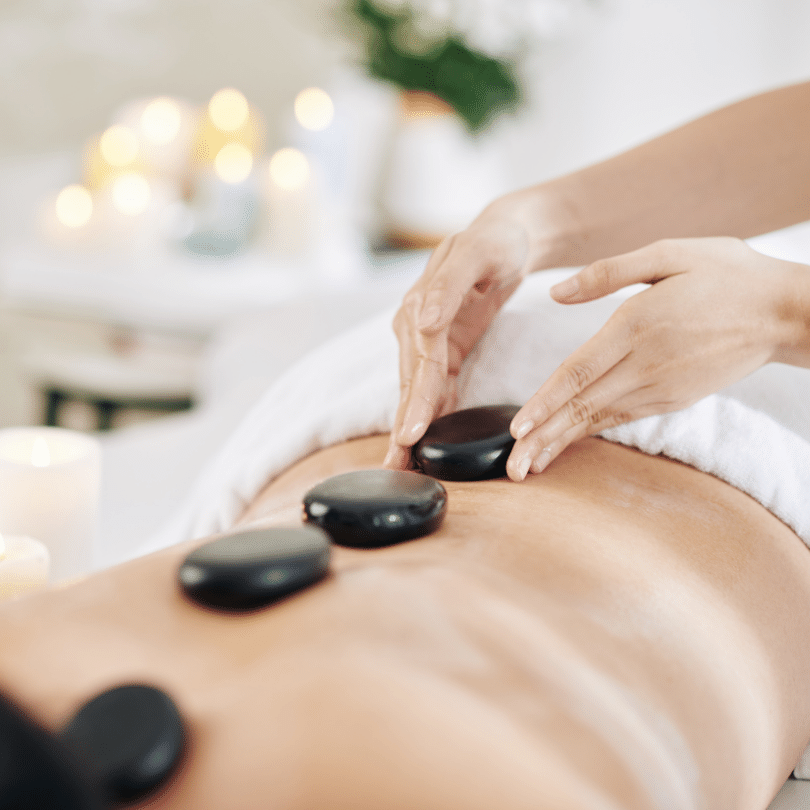 A tranquil hot stone massage session is beautifully captured in this image, where a client is seen relaxing on a massage table with a clean, white towel draped around the lower back. The therapist's skilled hands are in the process of placing heated, smooth, black stones on the client’s bare skin, targeting the muscles along the spine. The stones, known for their ability to retain heat and help soothe tight muscles, are contrasted against the client's soft skin tone. The background is softly focused, showcasing a serene and inviting massage room ambiance with glowing candles and plush decor, creating a sense of warmth and relaxation. This setting is ideal for promoting the therapeutic benefits of a hot stone massage, a practice celebrated for its relaxation and healing properties.