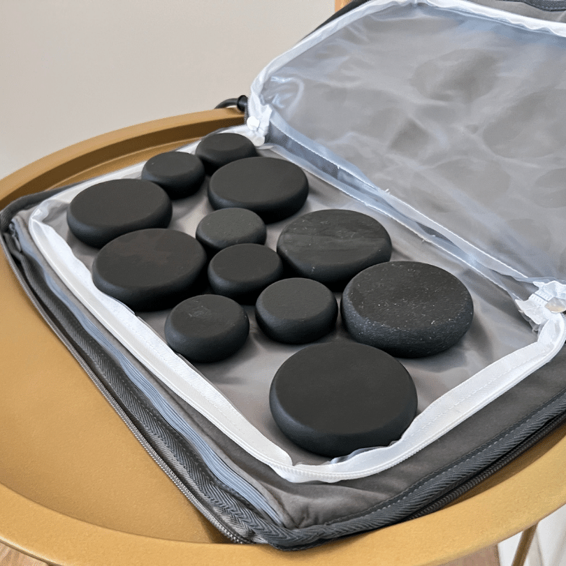 A collection of various-sized, smooth black hot stones prepared for a hot stone massage, neatly arranged in a specialized heating case to retain warmth. The stones are displayed on a soft, gray cushioned interior, contrasting their volcanic texture and deep color. This equipment is crucial for a hot stone massage, as the heated stones are used to relieve muscle tension and stress. The open case rests on a golden-yellow stool, indicating a spa or massage therapy setting where these stones will soon be used to provide therapeutic heat to a client's body. The image captures the essential tools before they are placed on the skin, highlighting the preparation and care taken in hot stone massage treatments to ensure a relaxing and beneficial experience for the client