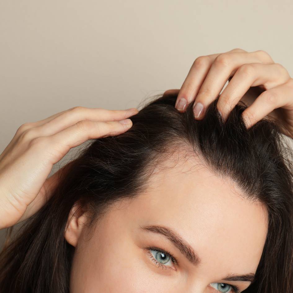 The image shows a close-up of a serene young woman with her eyes gently closed and a subtle smile on her lips, suggesting a moment of relaxation and stress relief, possibly after an enjoyable aromatherapy session in Burnaby. She's holding her hands up to her temples, lightly touching her face in a soothing manner. Her skin appears soft and clear, and she has neatly groomed eyebrows, natural makeup, and long eyelashes. The background is a plain, light color, emphasizing the calm and clean atmosphere. The woman's expression and gentle touch to her head convey a feeling of tranquility and self-care.